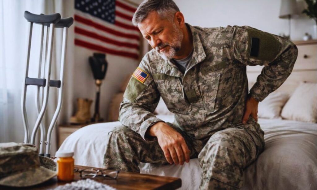 Disabled military veteran in uniform sitting on a bed beside crutches, reflecting on how a service related injury affects daily life.